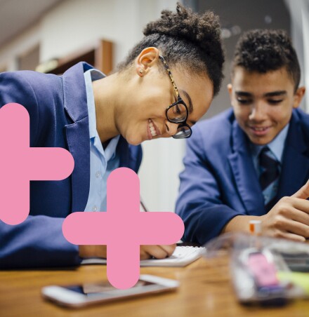 Secondary school kids are leaning on a desk and writing on a note pad. There are two pink coding symbols overlaid on the image to the left.