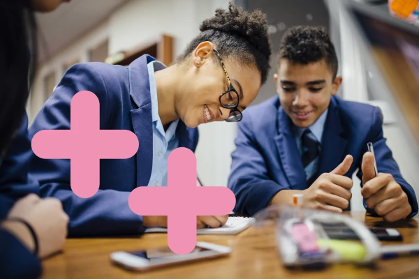 Secondary school kids are leaning on a desk and writing on a note pad. There are two pink coding symbols overlaid on the image to the left. 