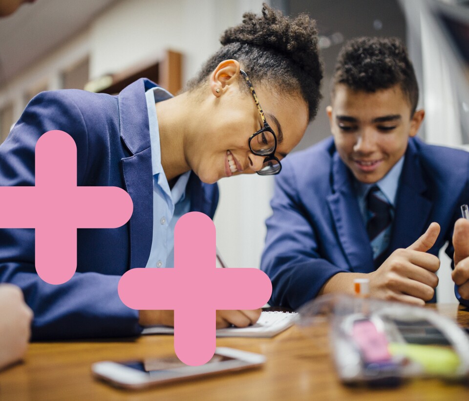 Secondary school kids are leaning on a desk and writing on a note pad. There are two pink coding symbols overlaid on the image to the left.