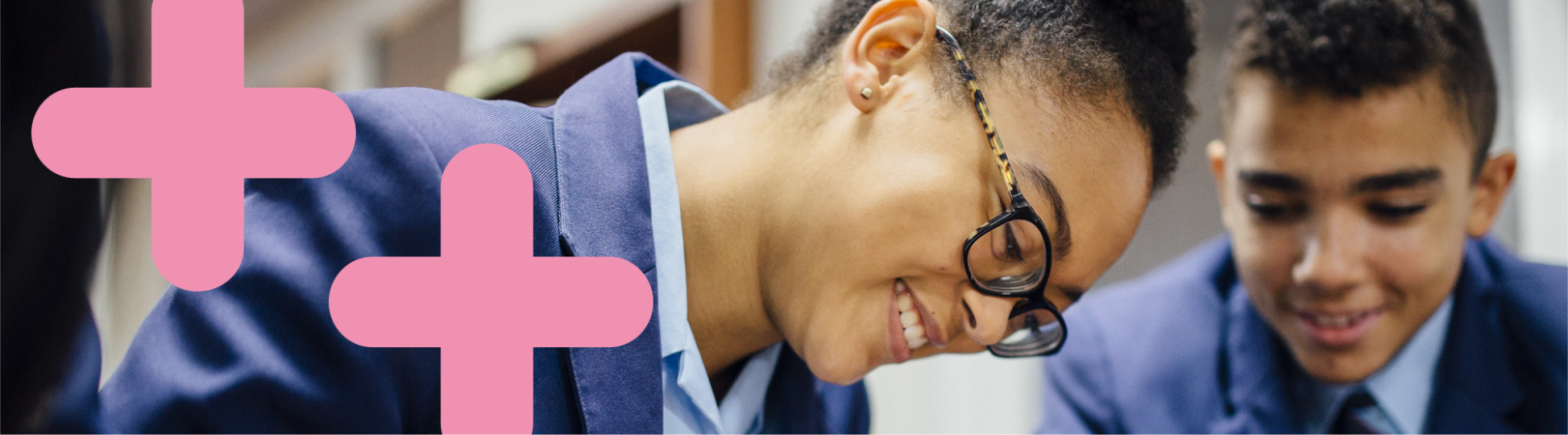 Two students work together and are smiling down at their work. There are two pink coding symbol graphics overlaid on the image to the left of the students.