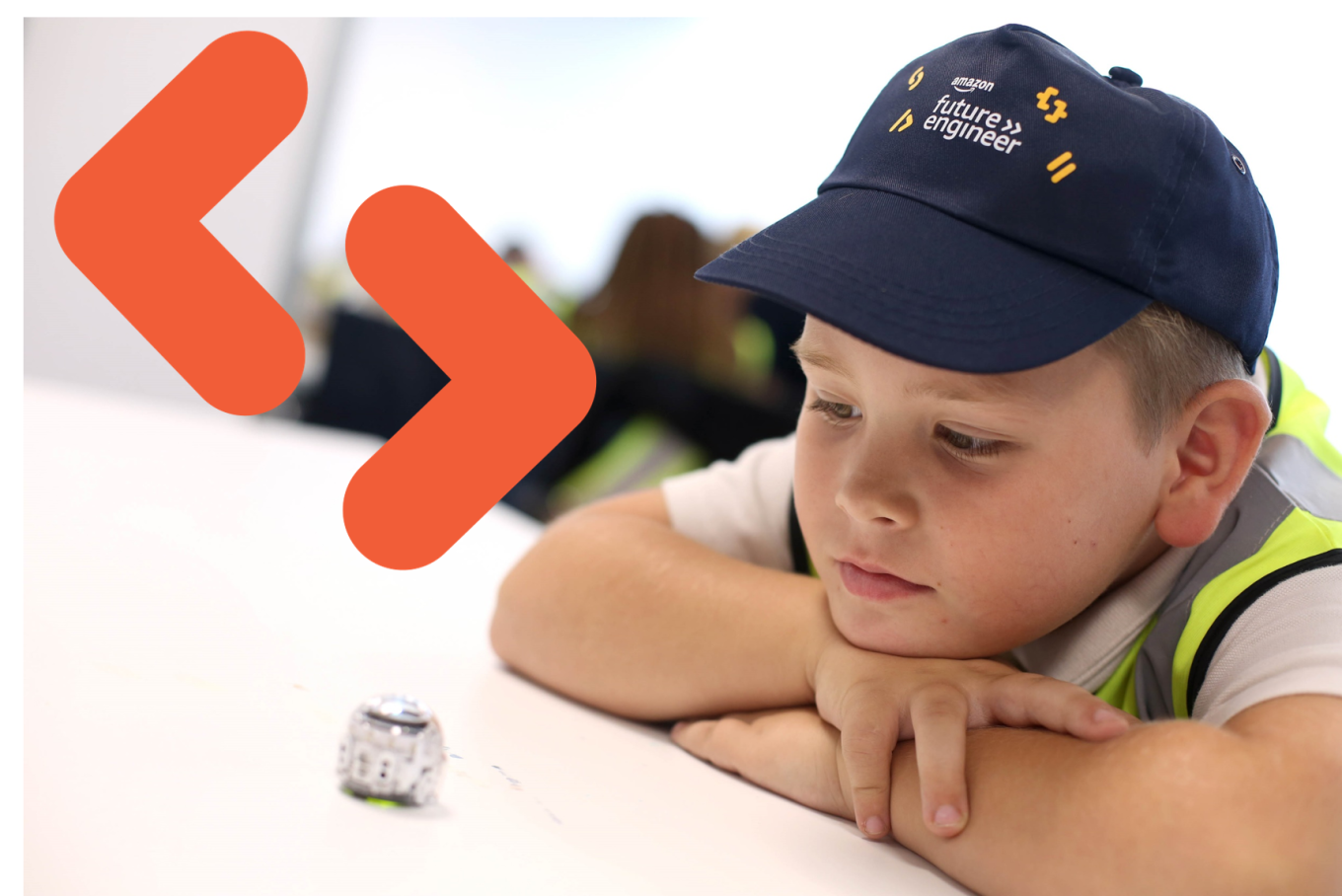 A primary student wearing an Amazon Future Engineer hat leans against a desk and looks down at a small device.